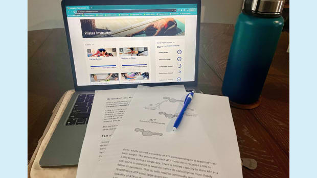 Study setup for ISSA Pilates Instructor course, with a lesson on a laptop, printouts for studying and a pen with a blue water bottle to the right of the computer